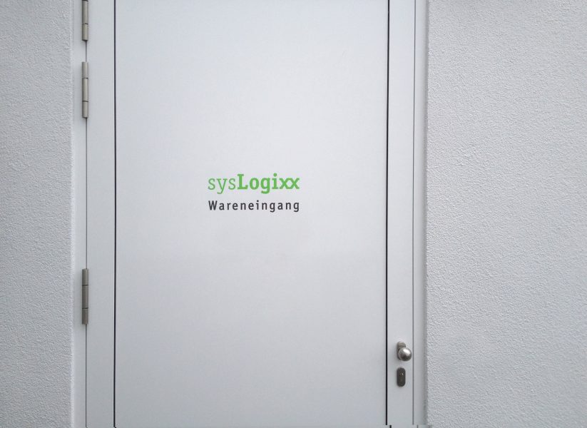Grünes sysLogixx Logo auf weißer Holztüre mit schwarzem Wareneingang Schriftzug