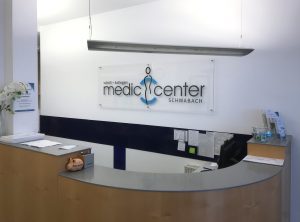 Transparentes Acrylschild mit dem Logo vom Medic Center an einer weißen Wand hinter dem Empfang