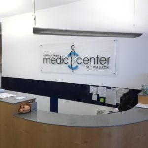 Transparentes Acrylschild mit dem Logo vom Medic Center an einer weißen Wand hinter dem Empfang