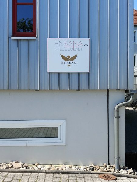 Ein foliertes Firmenschild aus Alu-Dibond an einer Außenwand dient als Wegweiser für den Ensana Pflegedienst und El Xeno