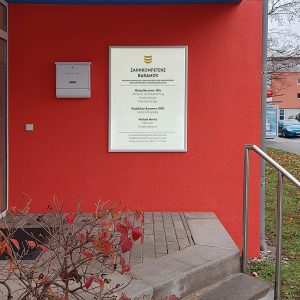 Ein Leuchtschild, das an der rot angestrichenen Hauswand im Eingang der Zahnarztpraxis Baramov hängt
